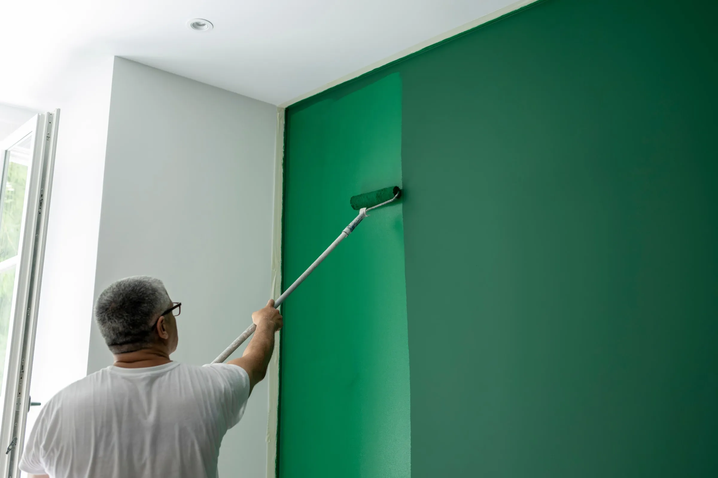 A painter rolling deep green paint onto an interior wall beside a bright window