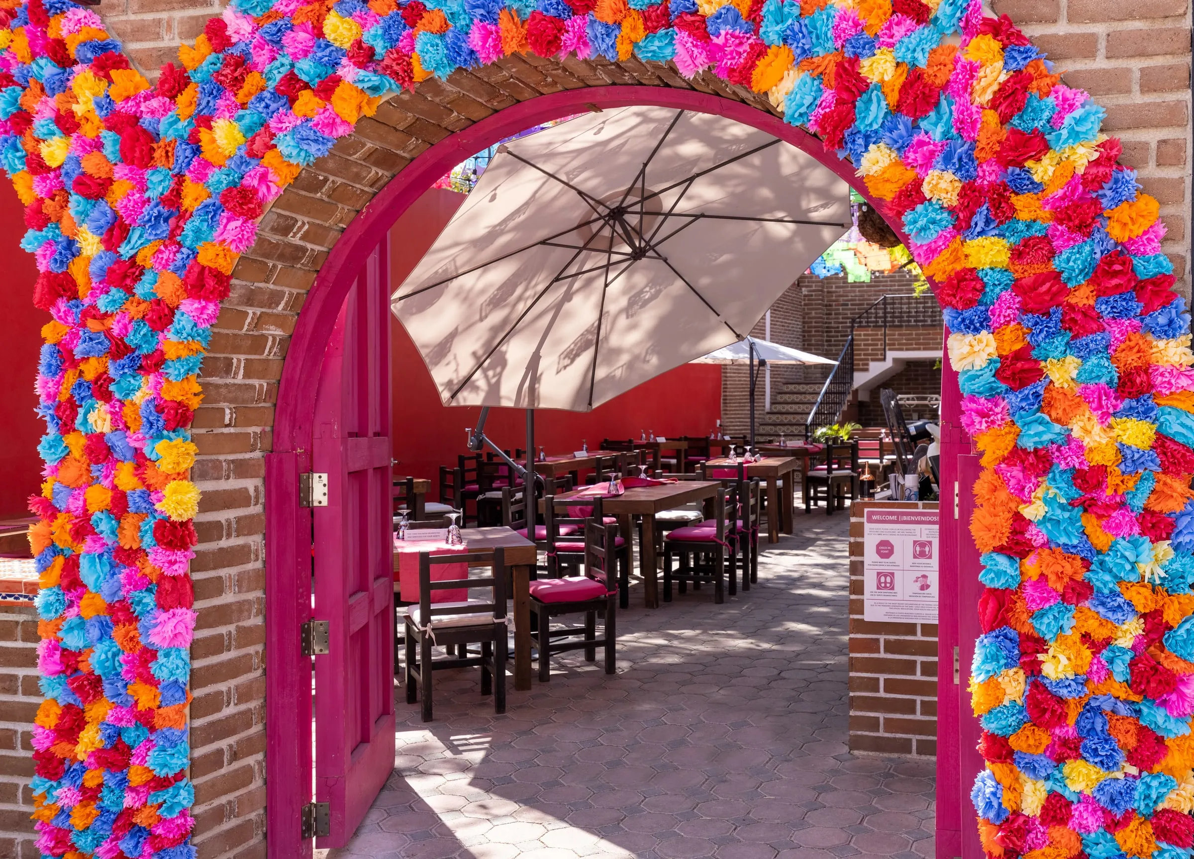 A brick archway framed in bright paper flowers, opening onto a restaurant patio of wooden tables under a parasol